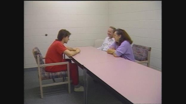 A young man in an orange jumpsuit sites at a table with a middle-aged woman and man sitting across from him.